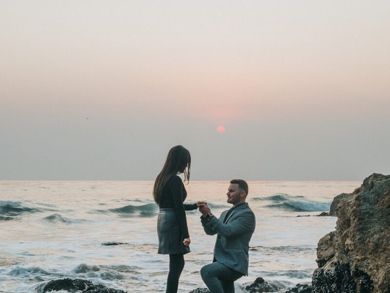 man kneeling in front of woman on rock at beach